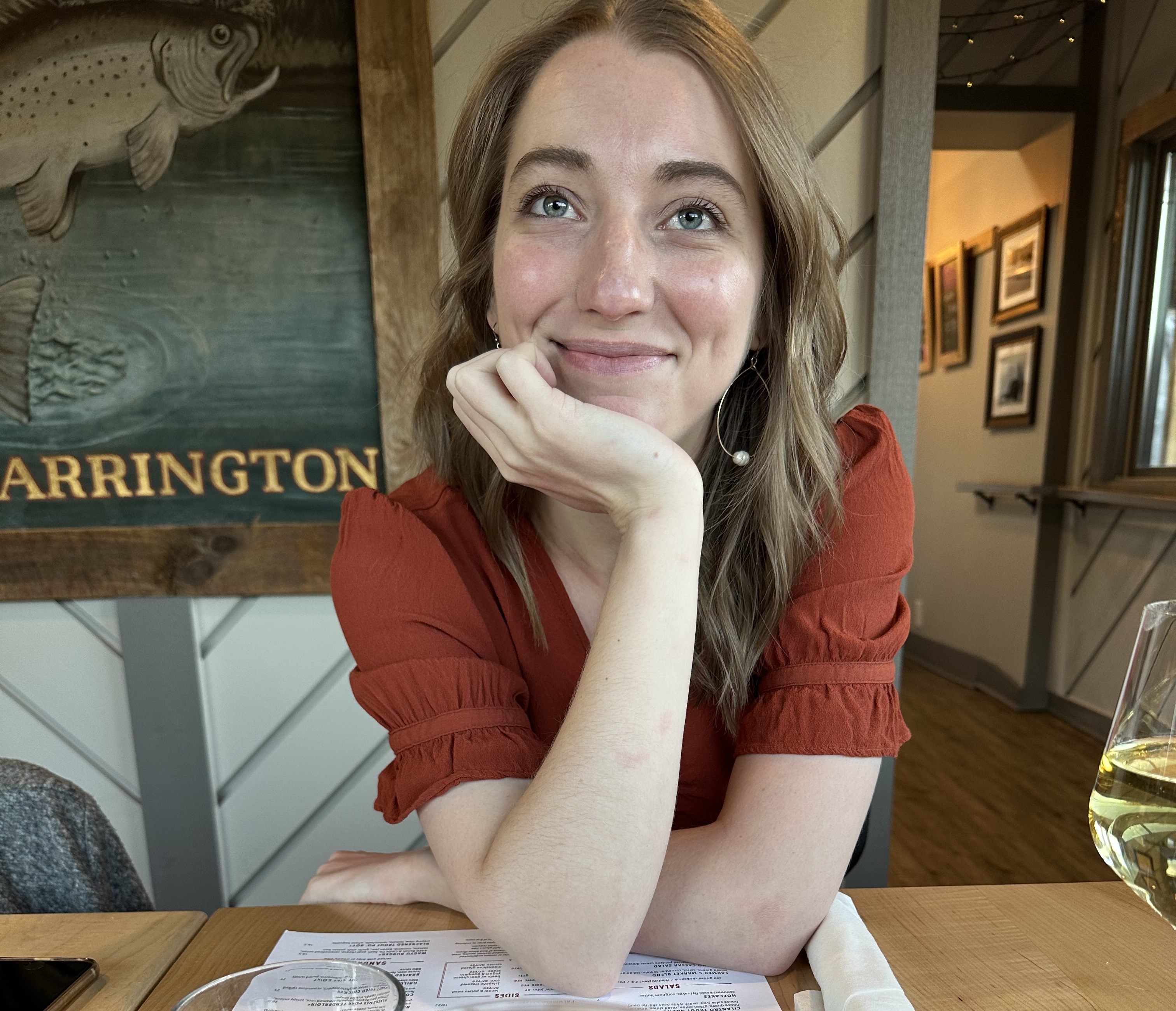 Britt Kreuger is a White woman sitting at a wooden table wearing a red/orange top.