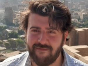 Tariq is a White man with dark hair wearing a blue checked shirt standing against an urban setting. 
