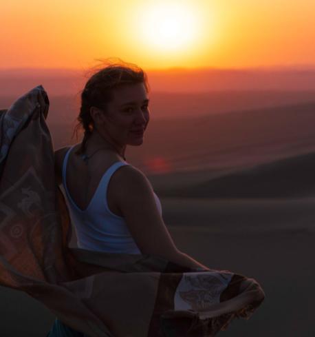 Anastasia Dorenbusch is a woman with dark hair wearing a white shirt. She stands in front of a sunset.