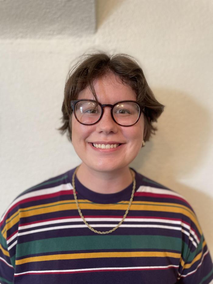 Lilly stands against a tan background. She is a White woman with short brown hair. Lilly is wearing a striped multi-color shirt and brown glasses.