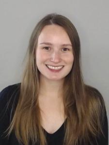 Rachel Lotze is a White woman with long brown hair. She is wearing a black shirt and stands against a grey background.