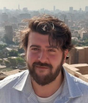 Tariq is a White man with dark hair wearing a blue checked shirt standing against an urban setting.